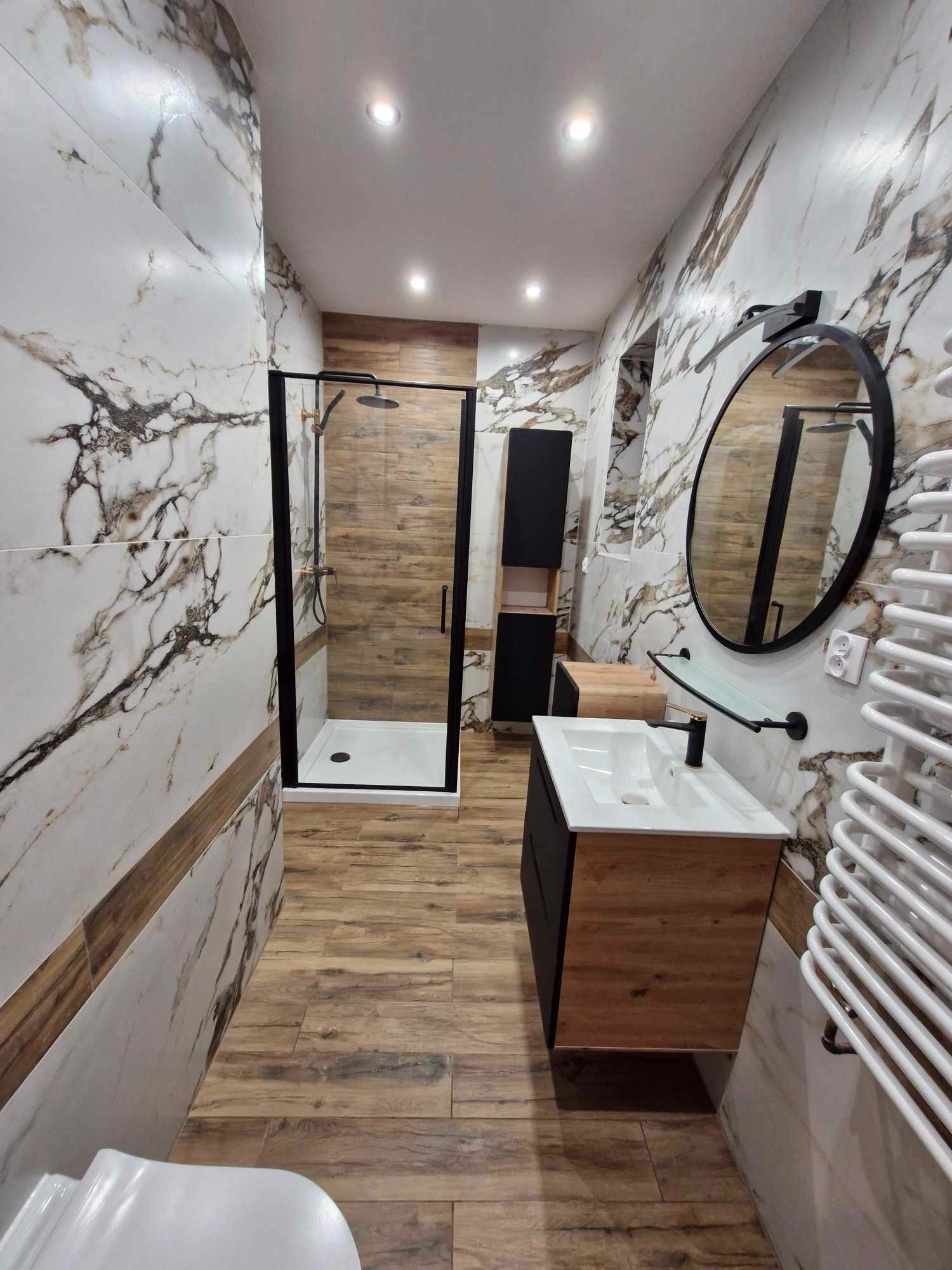 Nowoczesna łazienka z kabiną prysznicową z czarną ramą, umywalką z szafką w kolorze drewna i czarnymi elementami, marmurowe płytki na ścianach z drewnianym akcentem, drewnopodobna podłoga, okrągłe...