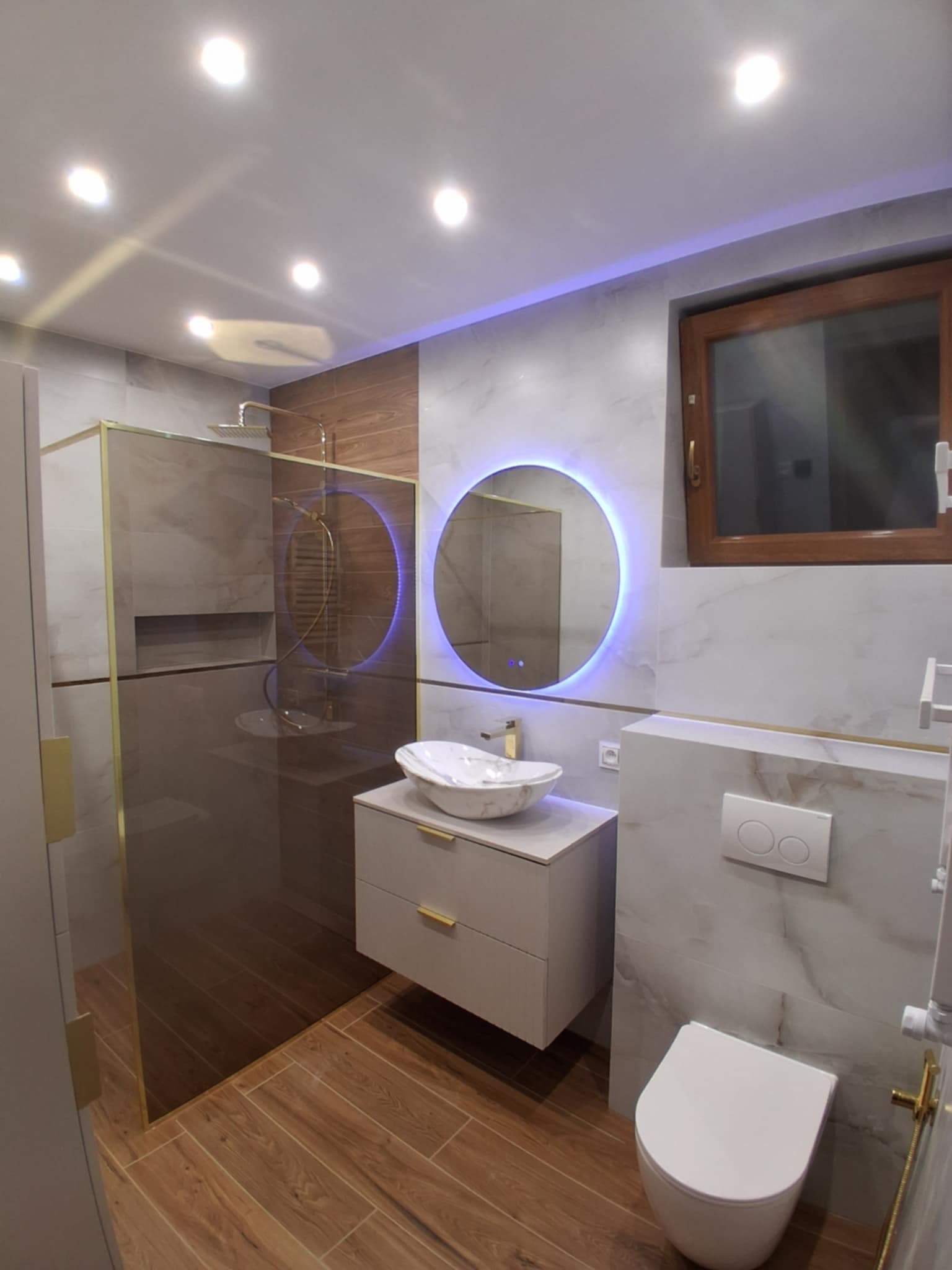 Nowoczesna łazienka po remoncie: marmurowe płytki, okrągłe lustro z podświetleniem LED, kabina prysznicowa ze złotymi detalami i drewnopodobna podłoga.