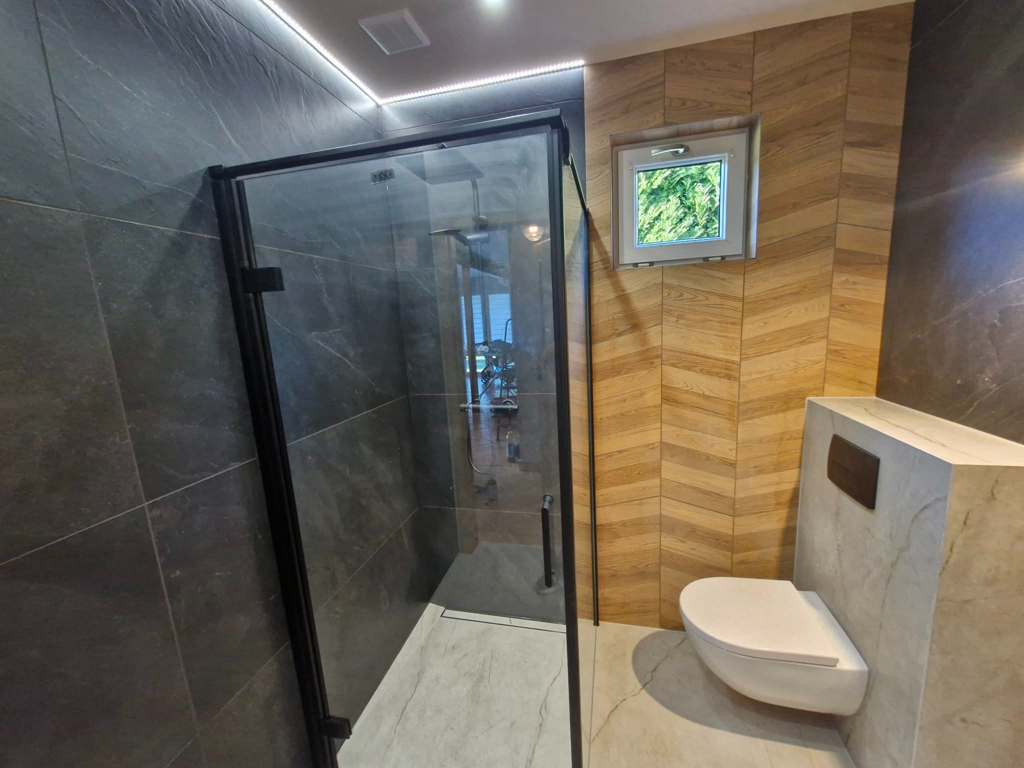 Nowoczesna łazienka z kabiną prysznicową typu walk-in z czarnymi profilami, drewnopodobnymi płytkami ułożonymi w jodełkę i podświetleniem LED. Wisząca toaleta.