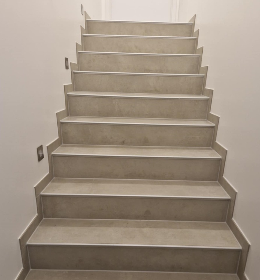 Wykończone schody wewnętrzne z beżowych płytek, oświetlenie punktowe na ścianie. Widok z dołu, symetryczny kadr.