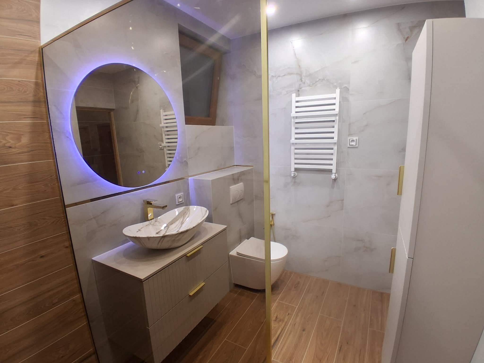 Wykończona łazienka z okrągłym lustrem podświetlanym na niebiesko, marmurową umywalką na szafce z złotymi uchwytami i grzejnikiem drabinkowym na ścianie z płytek imitujących marmur.