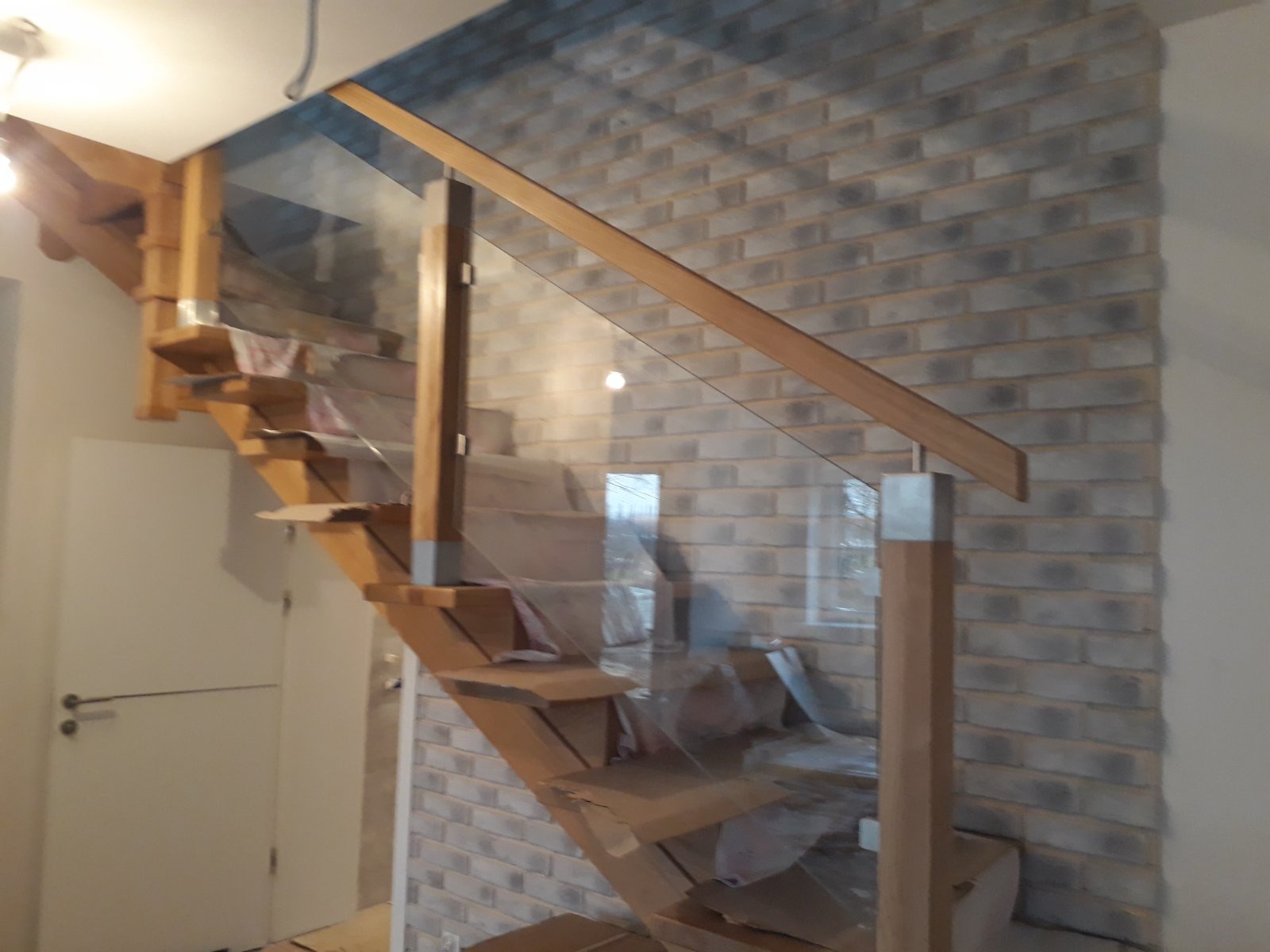 Drewniane schody z szklaną balustradą w trakcie montażu, zabezpieczone folią, na tle ściany z cegły dekoracyjnej w odcieniach szarości.