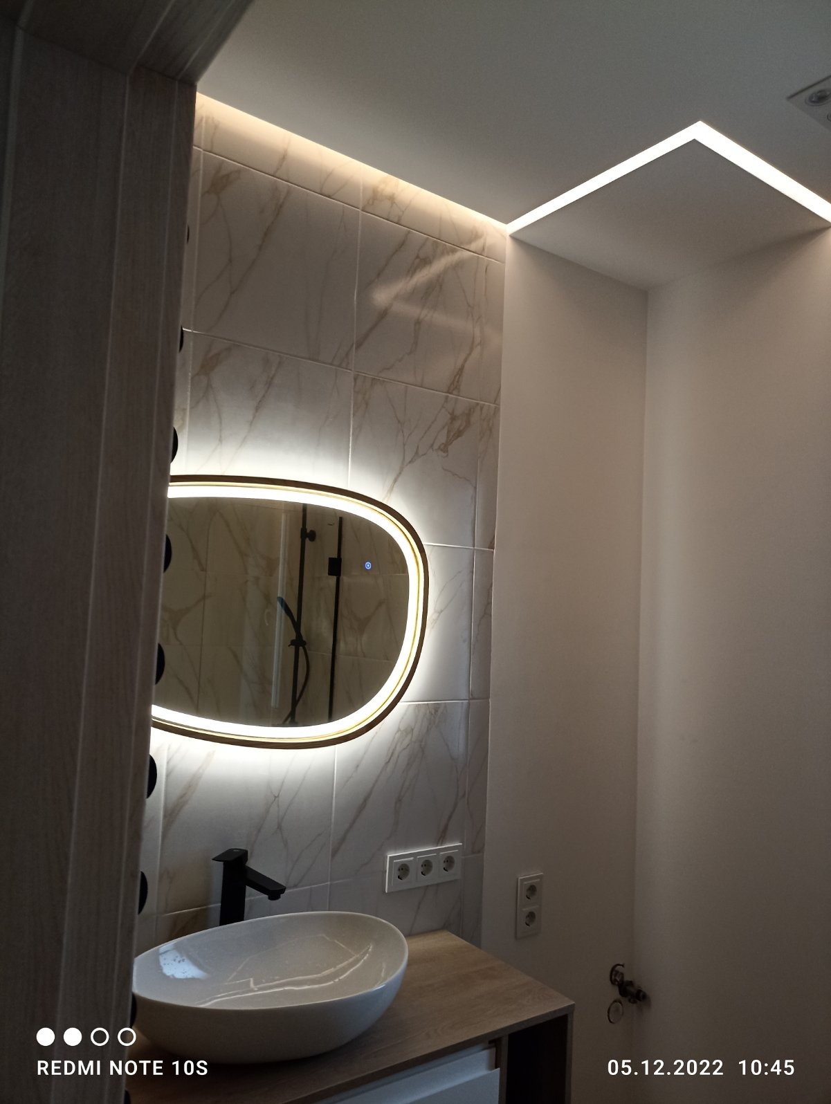 Nowoczesna łazienka z podświetlanym lustrem owalnym i marmurowymi płytkami, oświetleniem LED podsufitowym i białą umywalką nablatową na drewnopodobnym blacie.