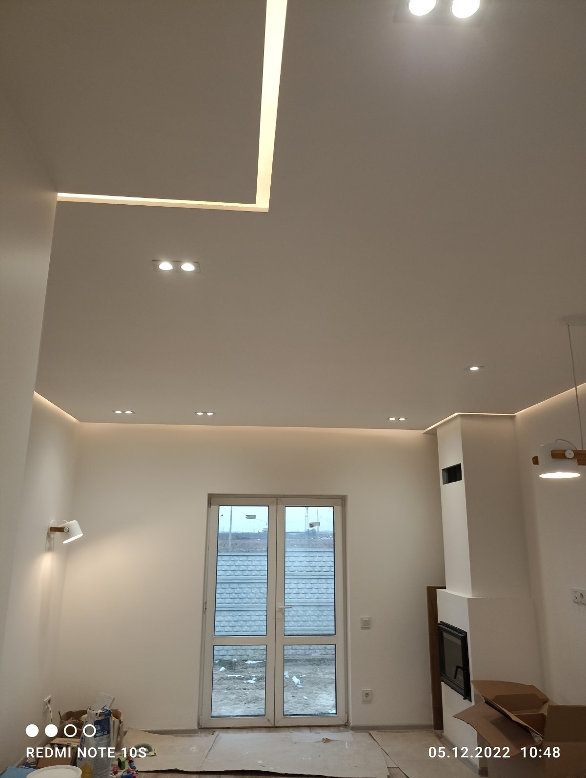 Nowoczesne oświetlenie sufitowe w salonie z kominkiem: listwy LED wpuszczone w sufit podwieszany, punktowe lampy sufitowe, białe ściany, widok na okno tarasowe i częściowo zakryte podłogi podczas...