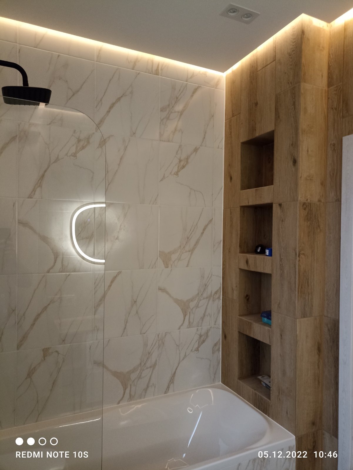 Wykończona łazienka w Skowarczu z białymi płytkami imitującymi marmur, podświetleniem LED i drewnianą zabudową z półkami wnękowymi.