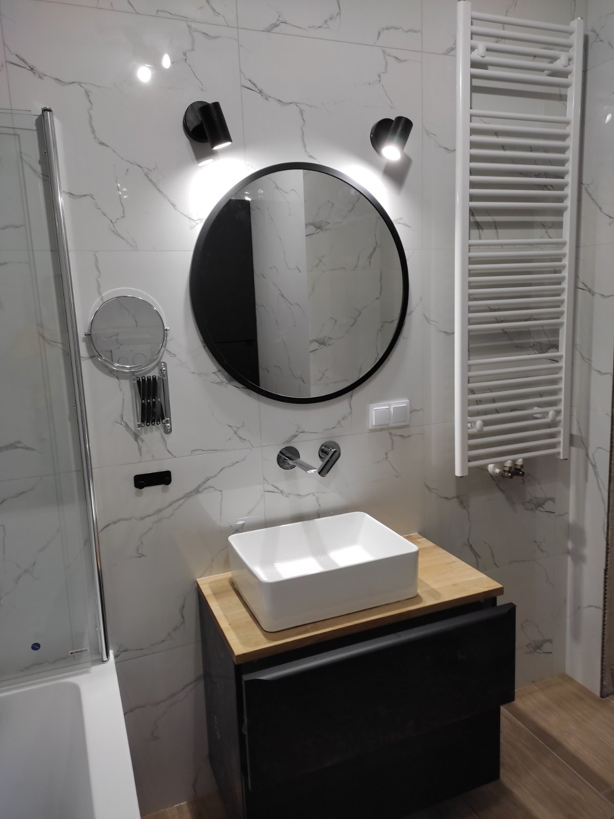 Nowoczesna łazienka z umywalką nablatową na szafce w kolorze antracytowym, okrągłym lustrem z czarną ramą, białymi płytkami imitującymi marmur oraz grzejnikiem drabinkowym.