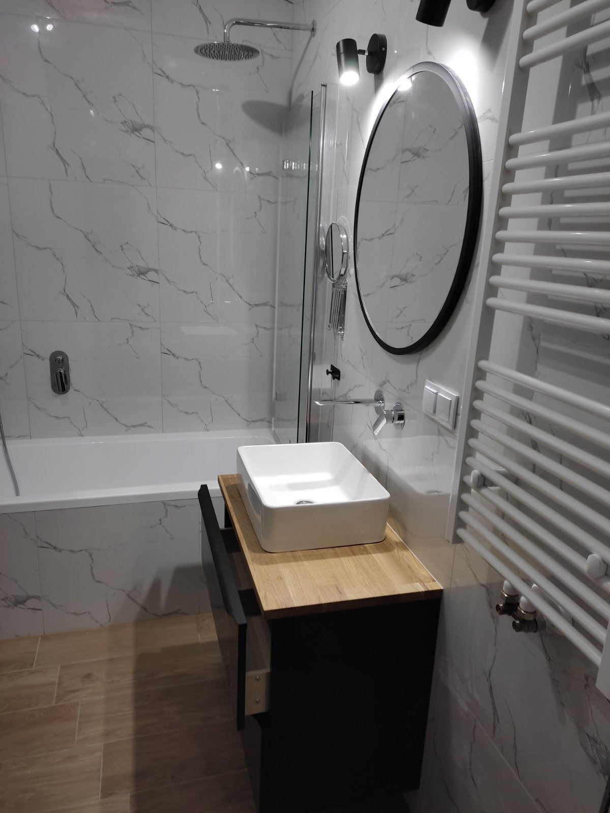 Nowoczesna łazienka z biało-szarymi marmurkowymi płytkami, owalnym lustrem w czarnej ramie, białą prostokątną umywalką nablatową na drewnianym blacie, grzejnikiem drabinkowym i kabiną prysznicową...