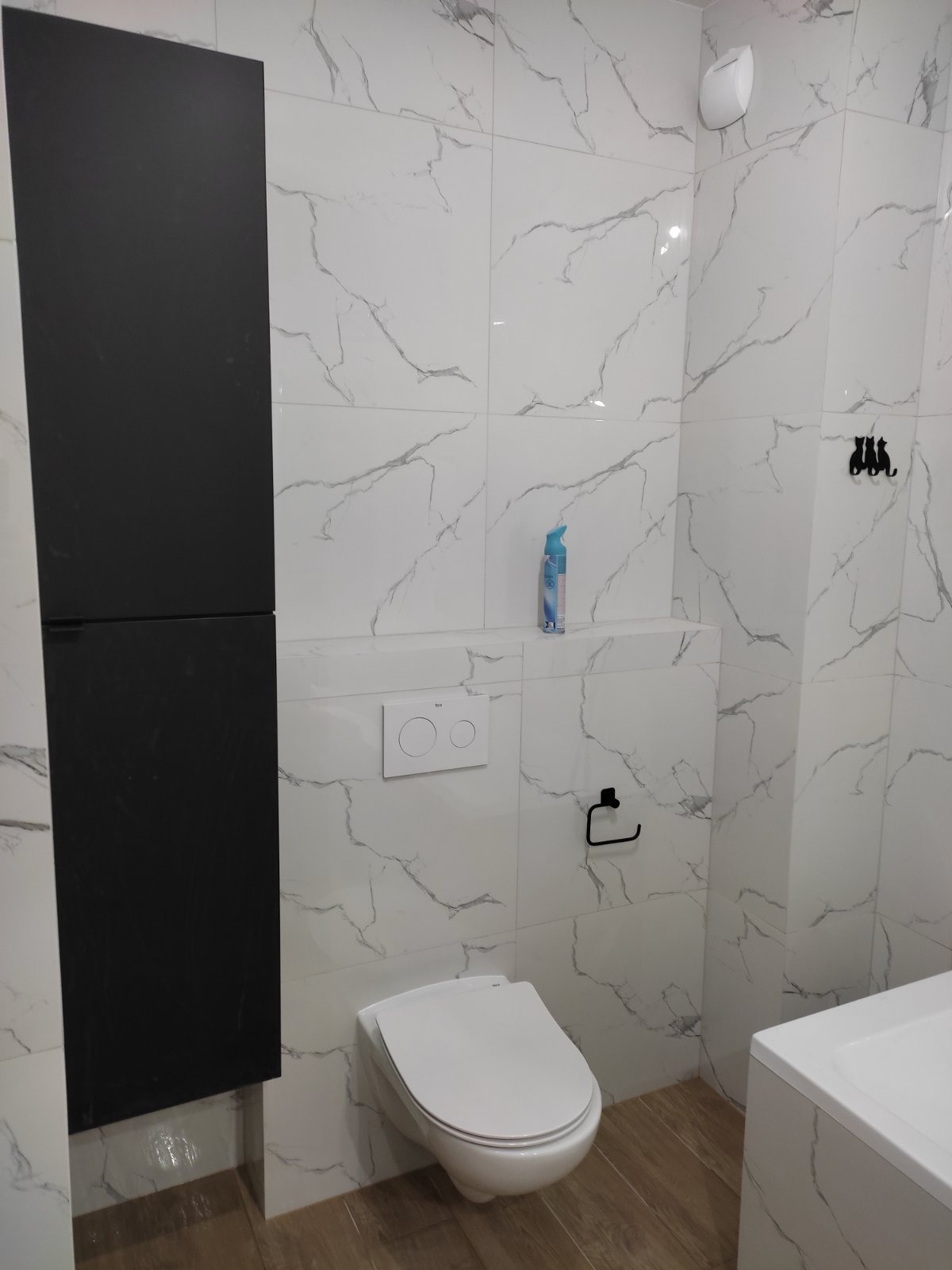 Nowoczesna łazienka z wiszącą toaletą, czarną szafką i marmurowymi płytkami z szarymi żyłami, drewnopodobna podłoga, designerski wieszak na ręczniki w kształcie trzech kotów.