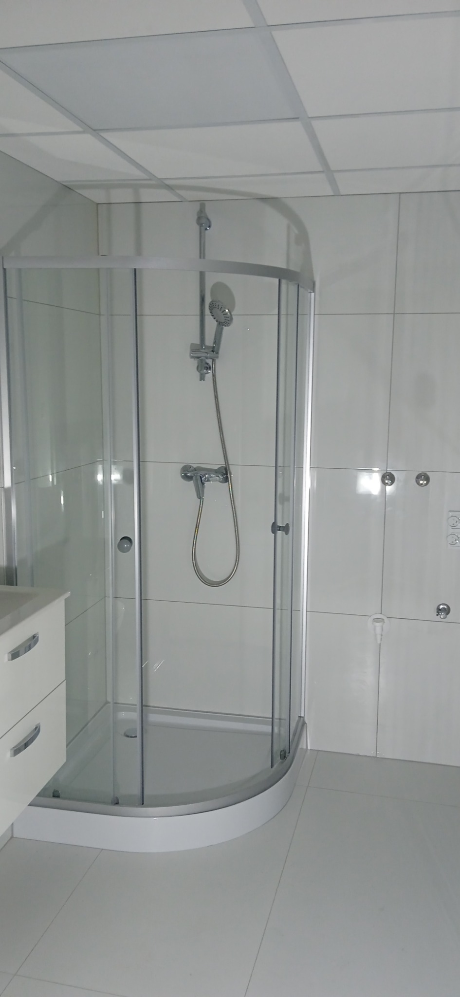 Nowoczesna kabina prysznicowa z zaokrąglonymi szybami i białą armaturą w łazience wykończonej białymi płytkami.