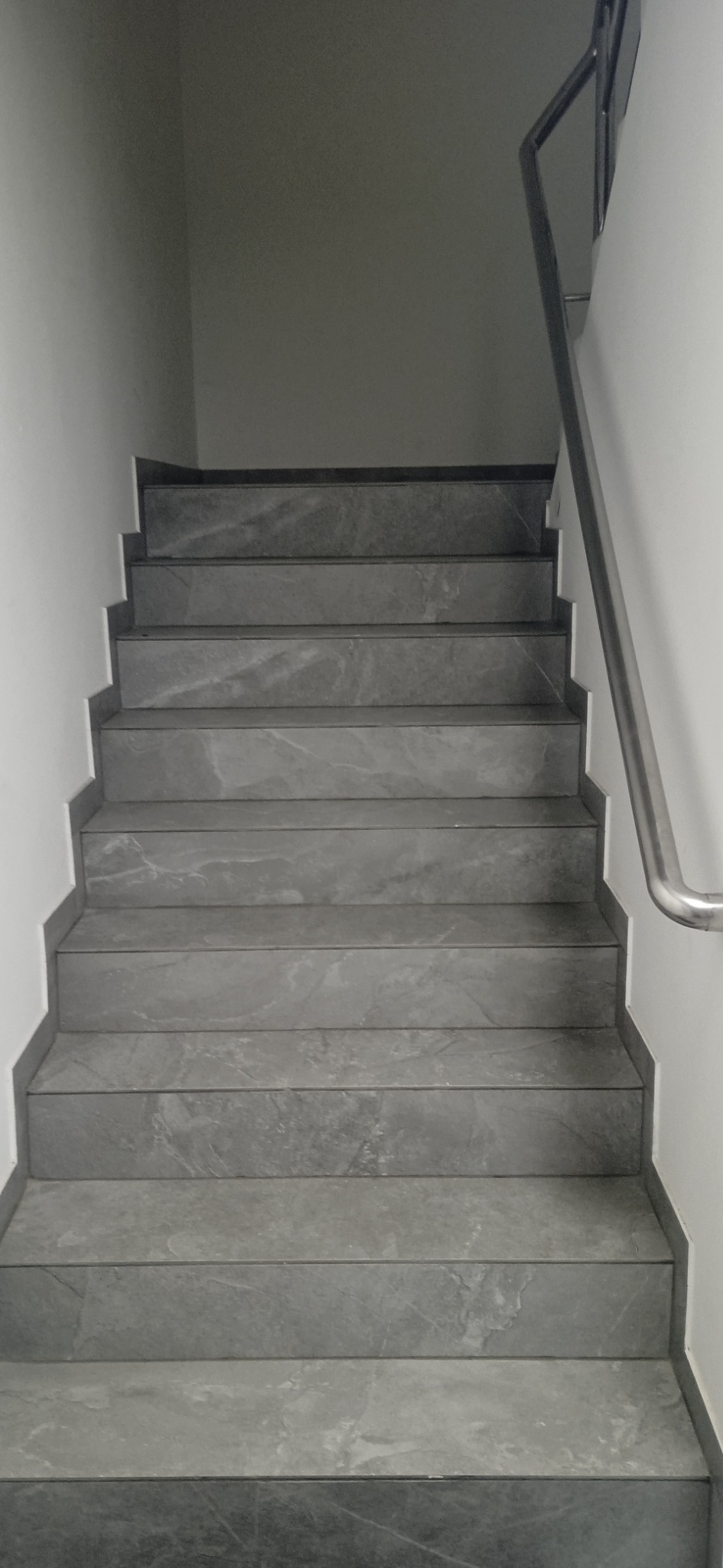 Schody wyłożone szarymi płytkami imitującymi kamień, z metalową poręczą po prawej stronie, widok z dołu.