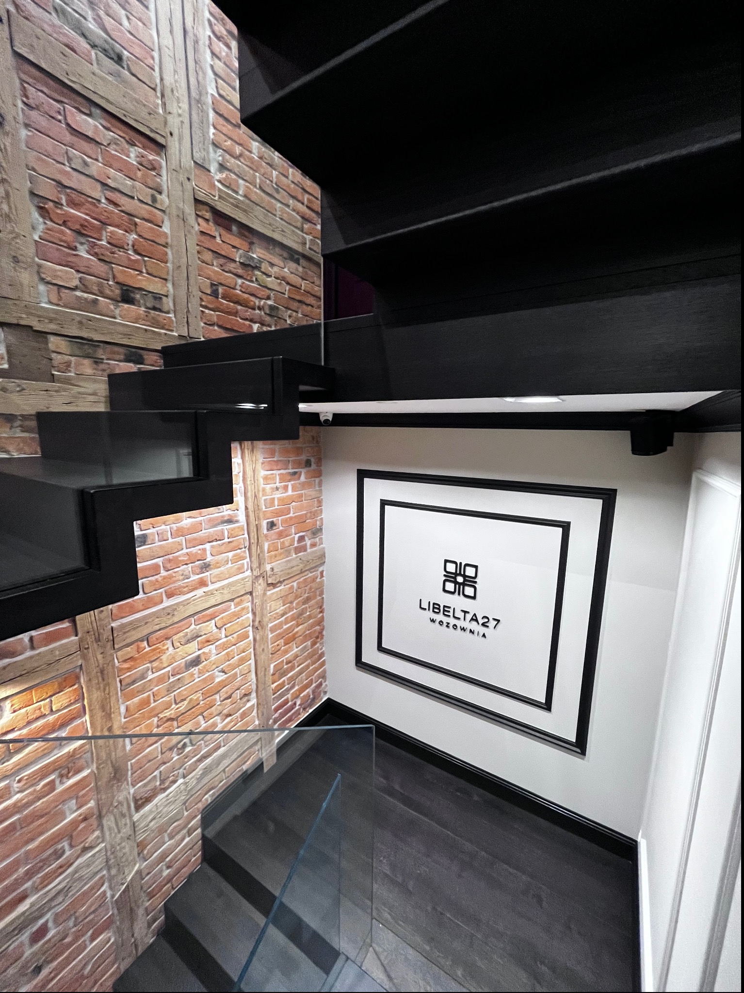 Nowoczesne, czarne schody z szklaną balustradą i ścianą imitującą cegłę, z obrazem firmy w czarnej ramie na białej ścianie.