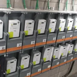 Rząd nowoczesnych liczników elektrycznych w szarych obudowach, zamontowanych na ścianie z szarej cegły, z widocznymi przewodami i oznaczeniami.