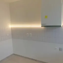 Nowoczesna kuchnia z białymi płytkami, podświetleniem LED pod szafkami i białym okapem z żółtą karteczką.