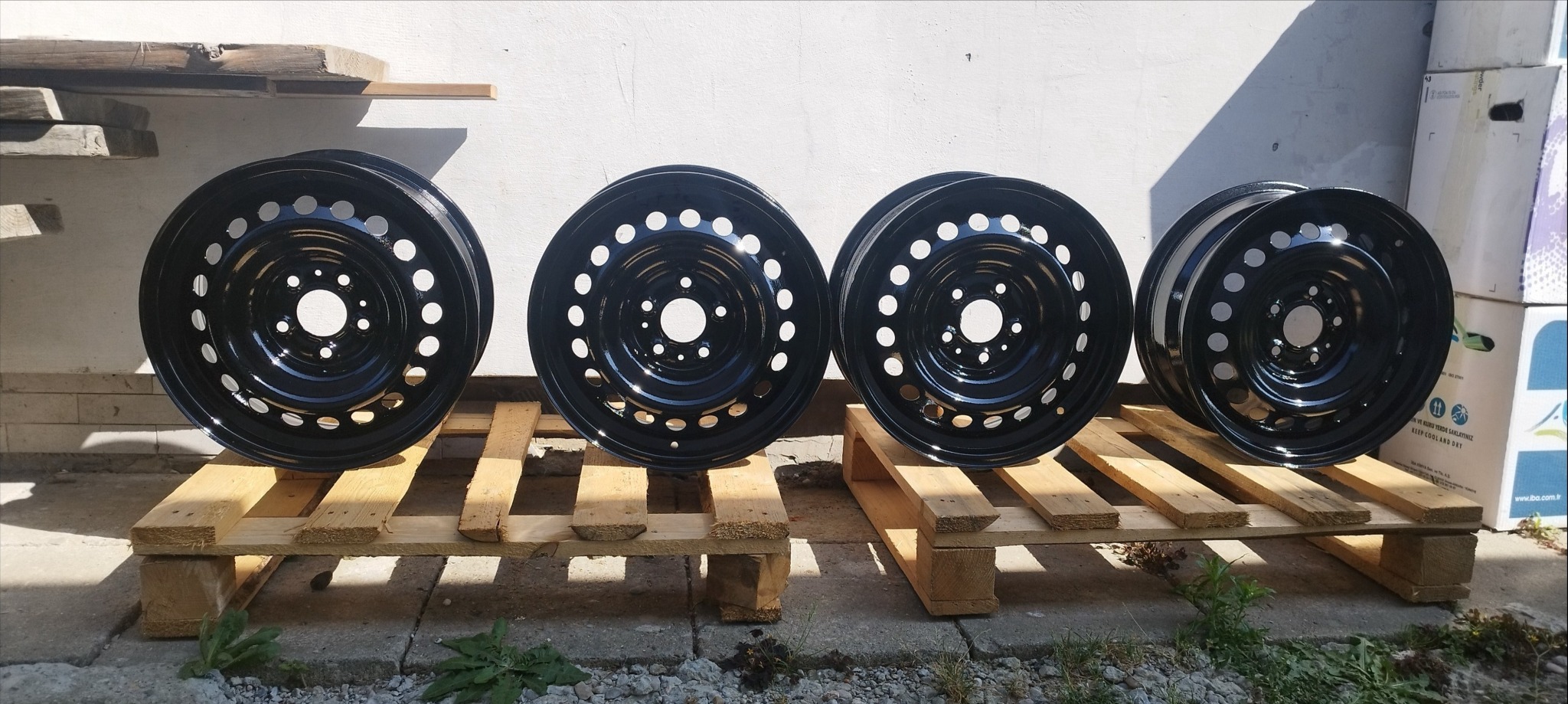 Cztery czarne, stalowe felgi samochodowe z otworami na palecie, po renowacji, z połyskiem, na tle białej ściany i kartonów.