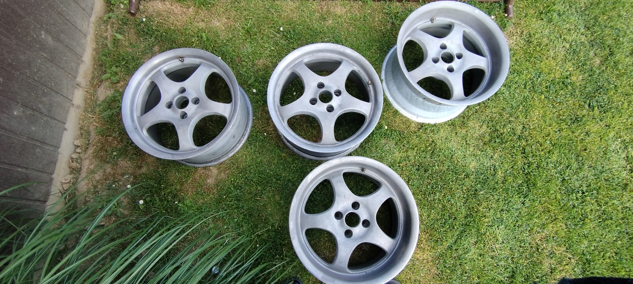 Cztery srebrne felgi samochodowe, leżące na trawie, po procesie oczyszczania, przygotowane do dalszej obróbki lub malowania.