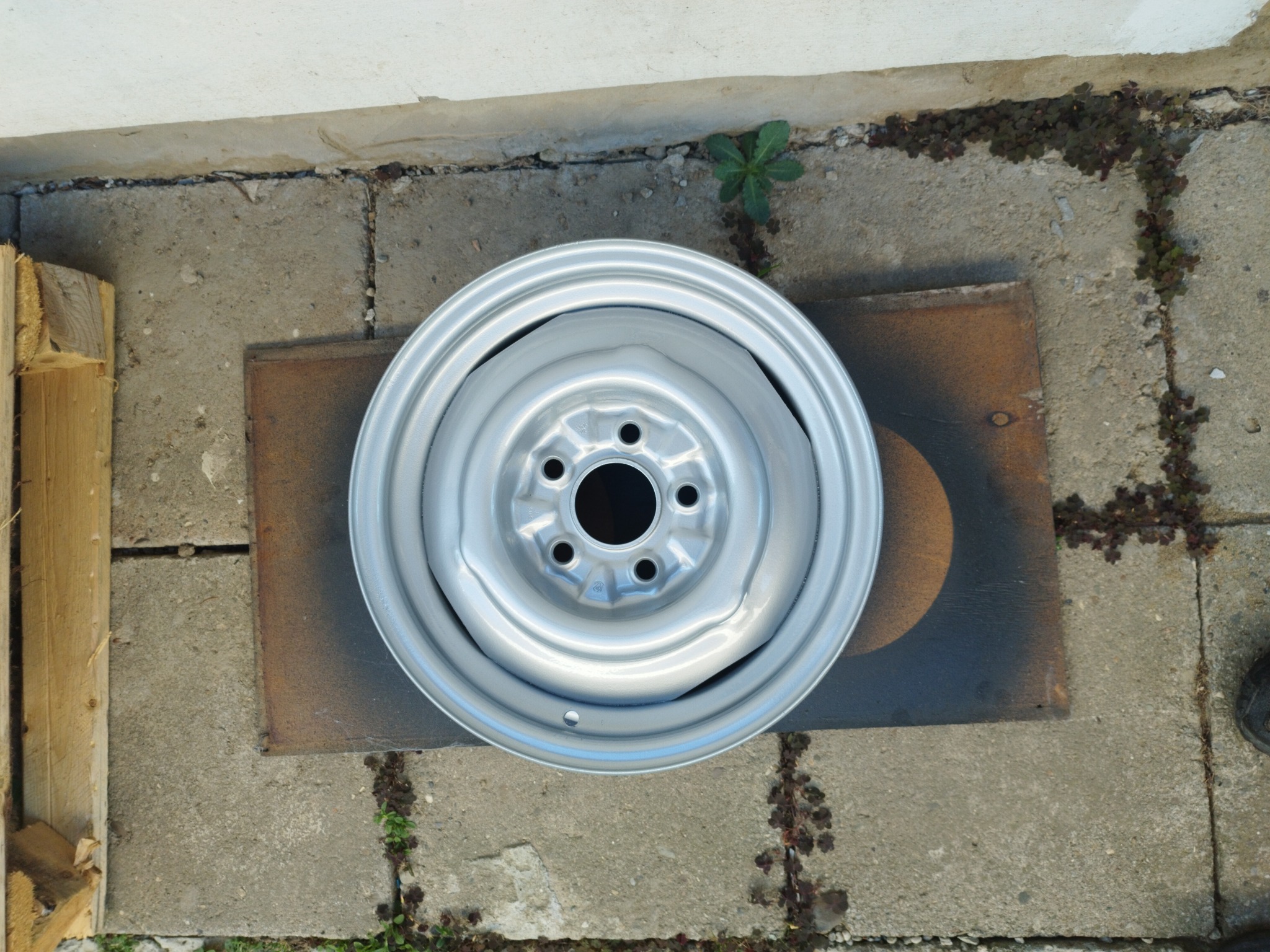 Pomalowana na srebrno felga samochodowa leży na brązowej blasze na betonowym podłożu z drobną zielenią.