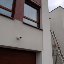 Biała kamera monitoringu zamontowana na elewacji budynku we Wrocławiu, obok brązowej bramy garażowej, z drabina opartą o ścianę w tle.