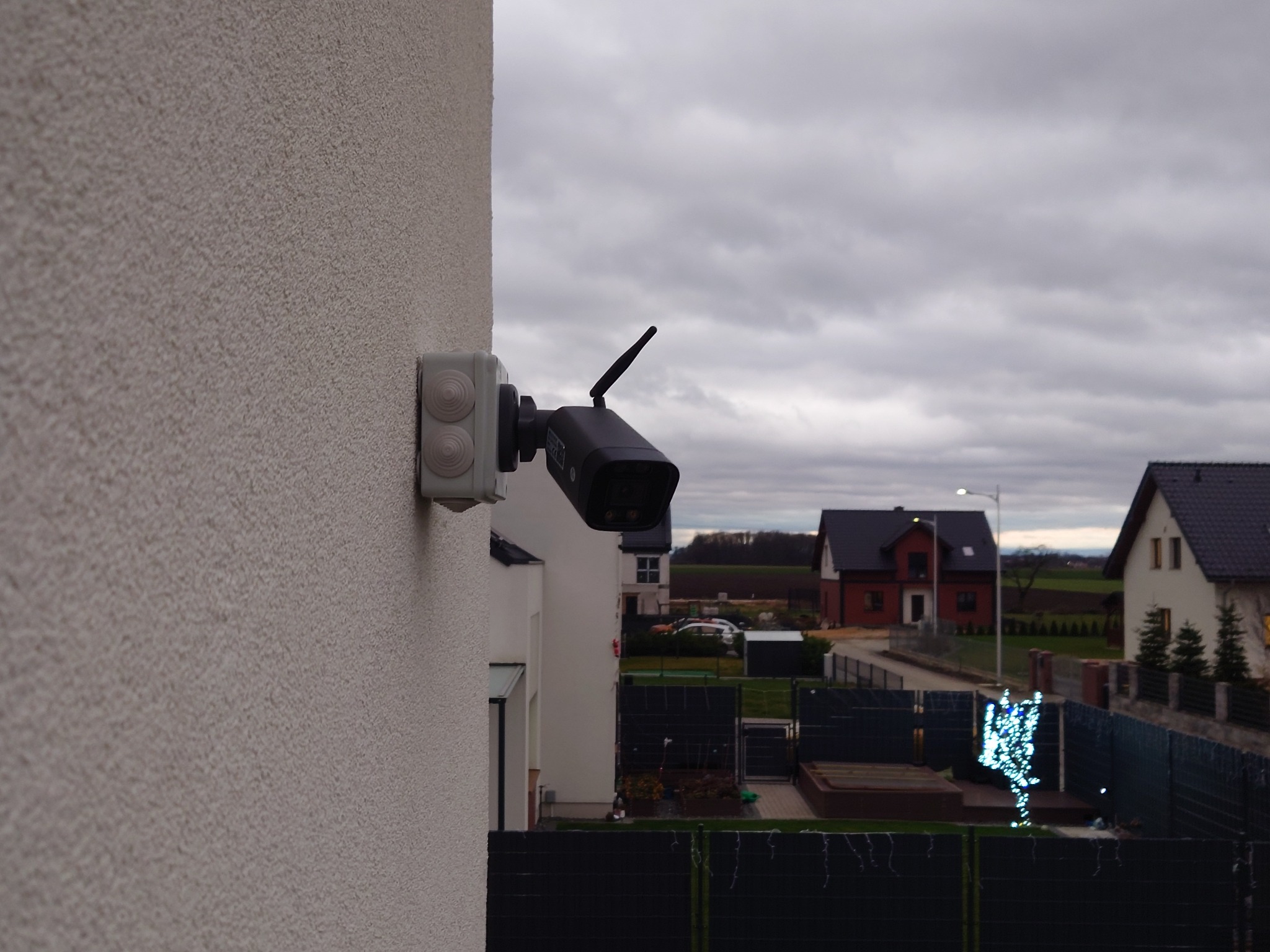 Zewnętrzna kamera monitoringu z anteną, zamontowana na elewacji domu, widok na sąsiednie posesje w pochmurny dzień.