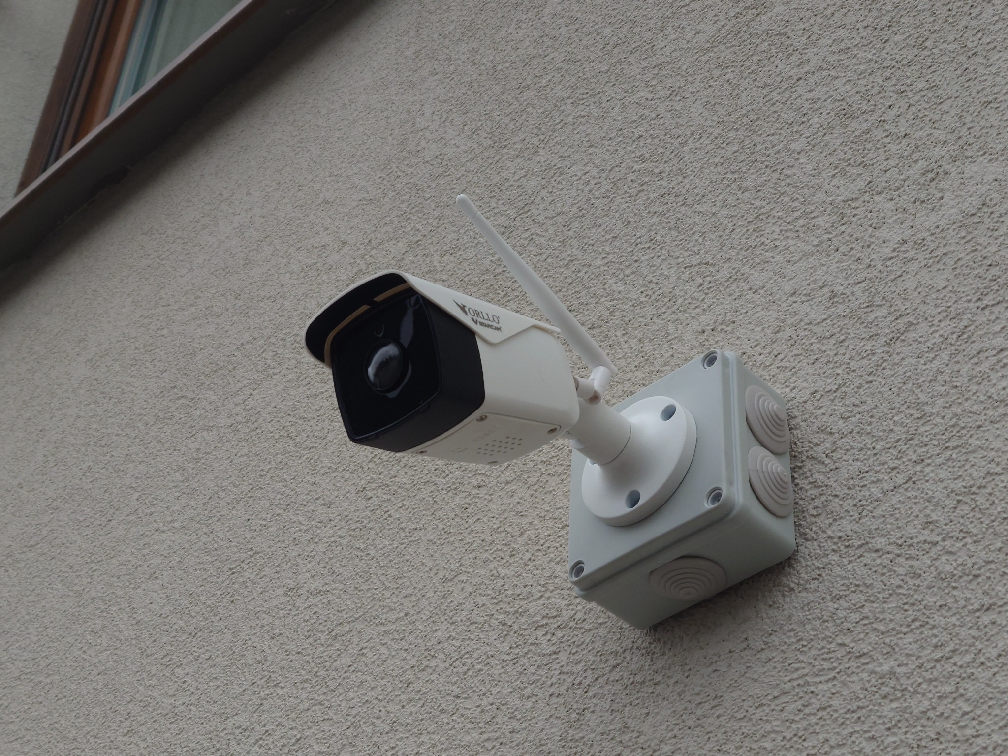 Zewnętrzna kamera monitoringu marki Orllo, zamontowana na jasnej, teksturowanej ścianie budynku, widoczna antena i puszka instalacyjna z gumowymi zaślepkami.