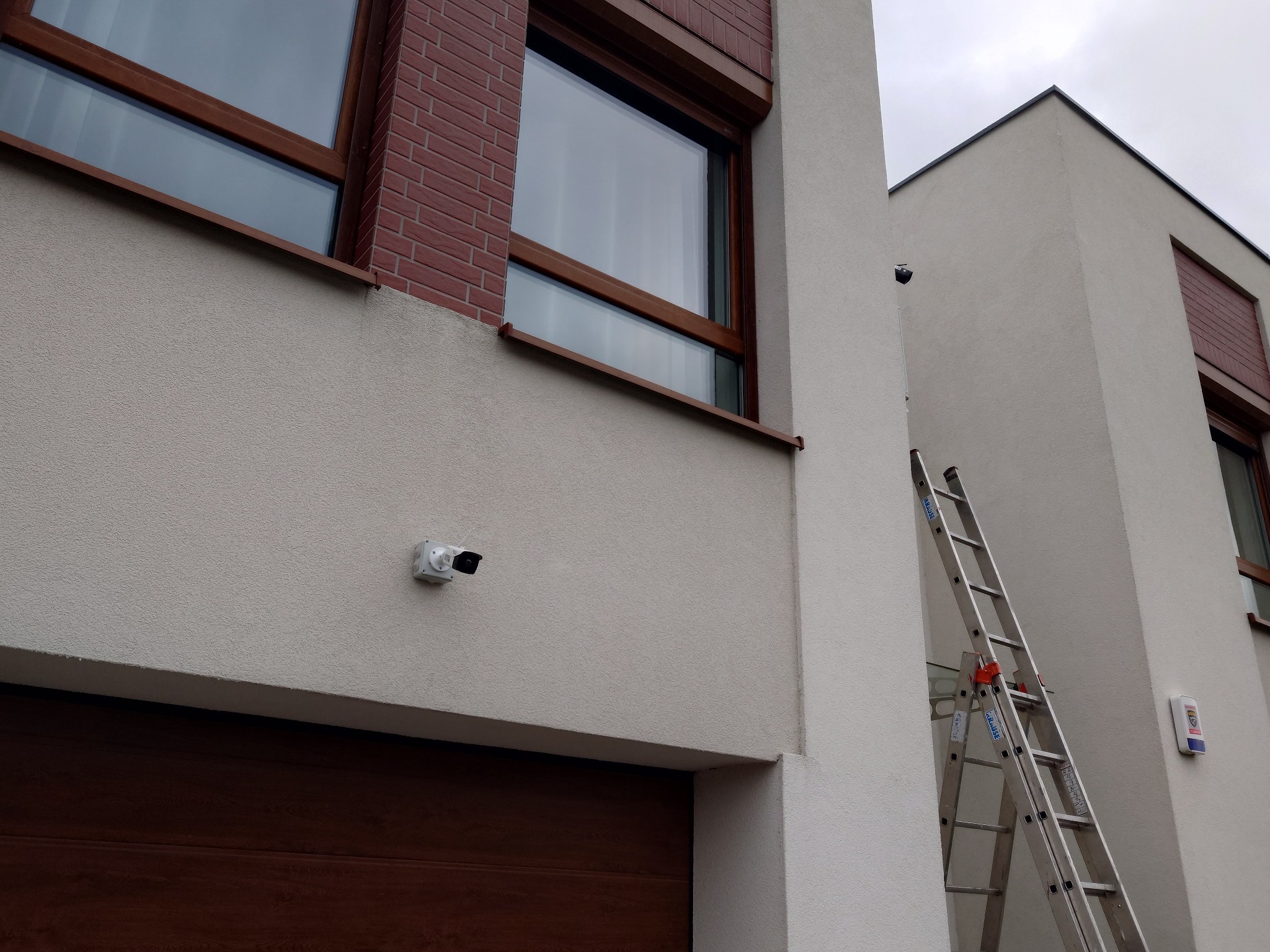Biała kamera monitoringu zamontowana na elewacji budynku we Wrocławiu, obok brązowej bramy garażowej, z drabina opartą o ścianę w tle.