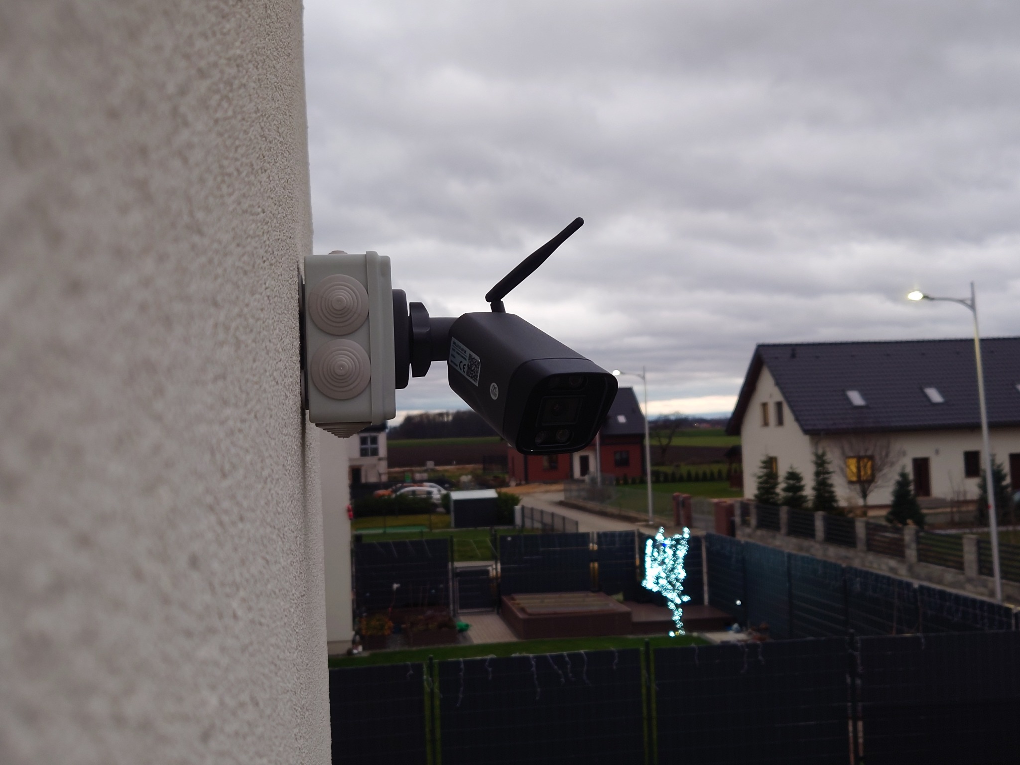 Zewnętrzna kamera monitoringu przymocowana do ściany budynku z widoczną anteną, z tłem przedstawiającym zabudowę jednorodzinną i pochmurne niebo.