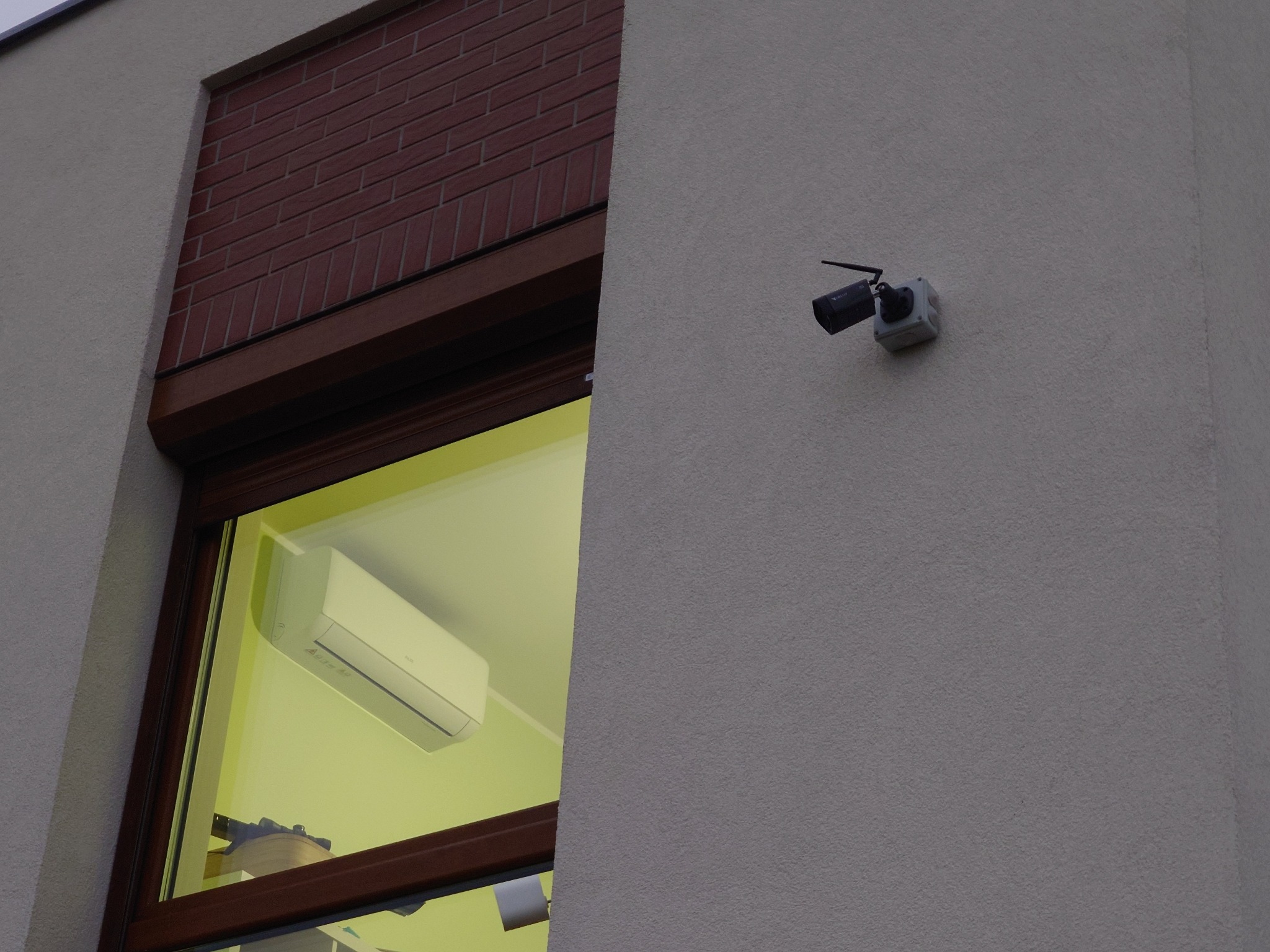 Zewnętrzna kamera monitoringu z antenką zamontowana na szarej, teksturowanej ścianie budynku, widoczna także część okna z brązowymi ramami i roletą oraz klimatyzator wewnątrz pomieszczenia.