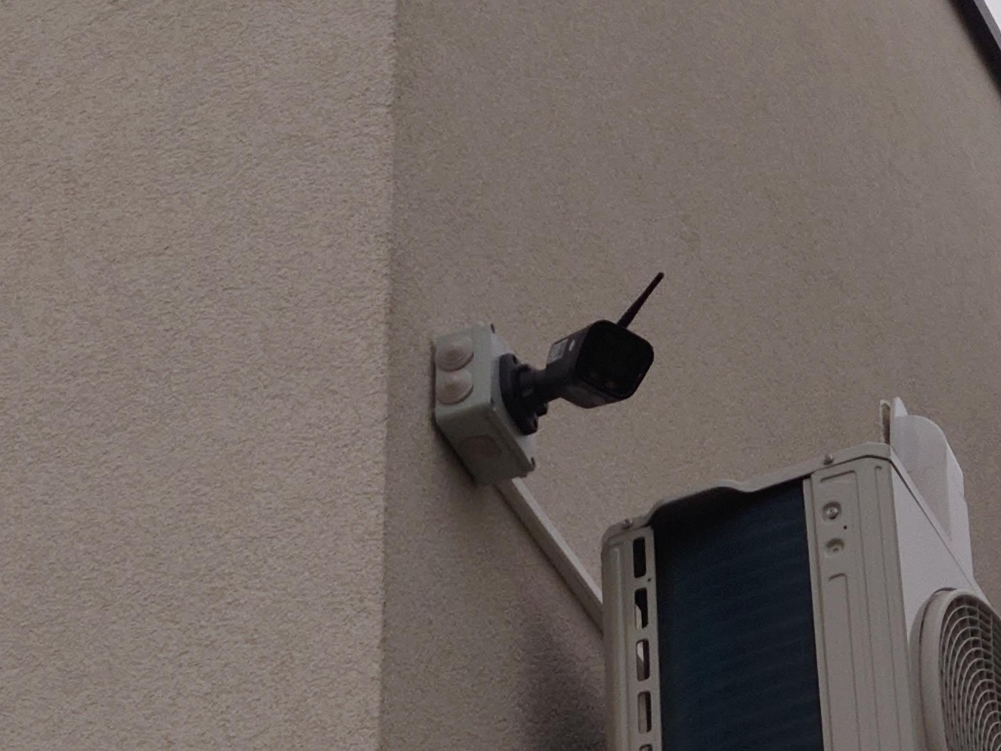 Zewnętrzna kamera monitoringu z anteną, zamontowana na elewacji budynku obok jednostki klimatyzacji.