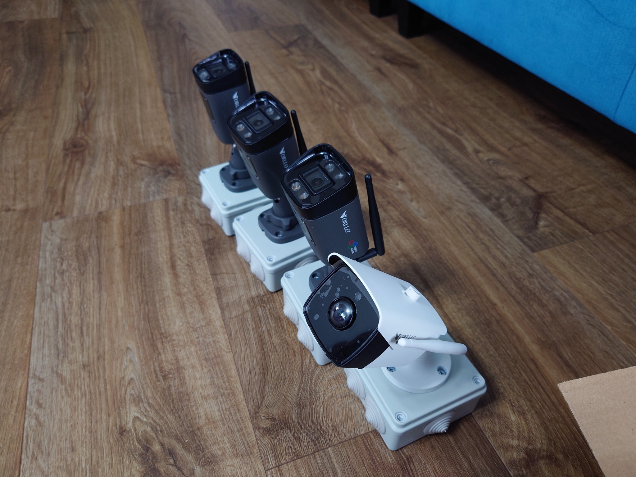 Cztery kamery monitoringu Orllo, trzy czarne i jedna biała, ustawione rzędem na drewnianej podłodze, z widocznym fragmentem niebieskiej sofy w tle.
