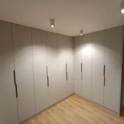 Jasnoszara szafa wnękowa z ciemnymi, pionowymi uchwytami, oświetlona punktowymi lampami sufitowymi, widok na parkiet z paneli.