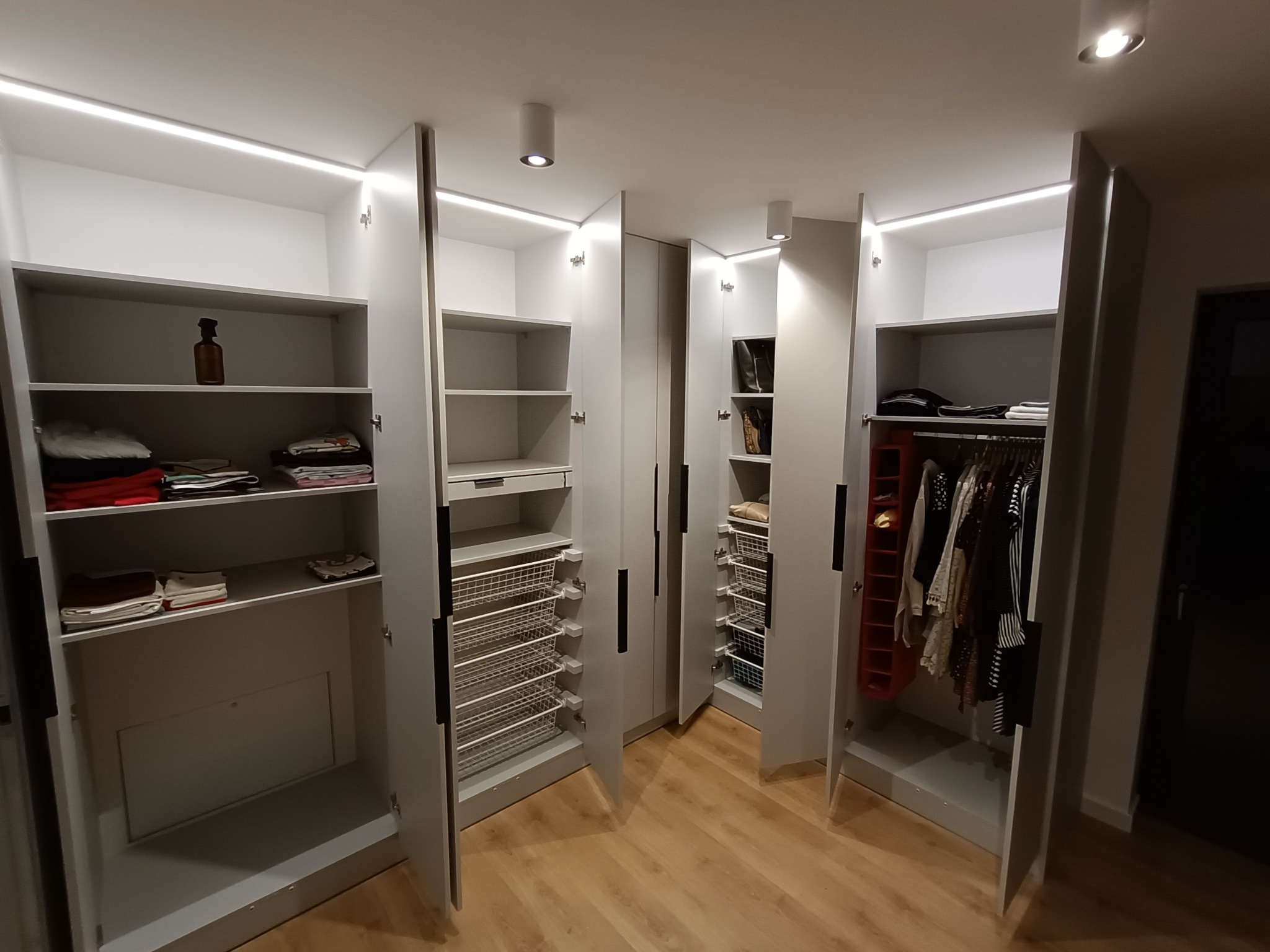Przestronna garderoba z białymi szafami z otwartymi drzwiami, oświetleniem LED podszafkowym i punktowym, prezentująca półki, wysuwane kosze i wieszaki z ubraniami.