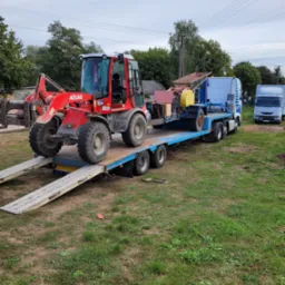 Czerwona ładowarka kołowa Atlas wjeżdża po rampie na niskopodwoziową naczepę ciężarówki, transportująca również inny sprzęt budowlany, na tle zieleni.