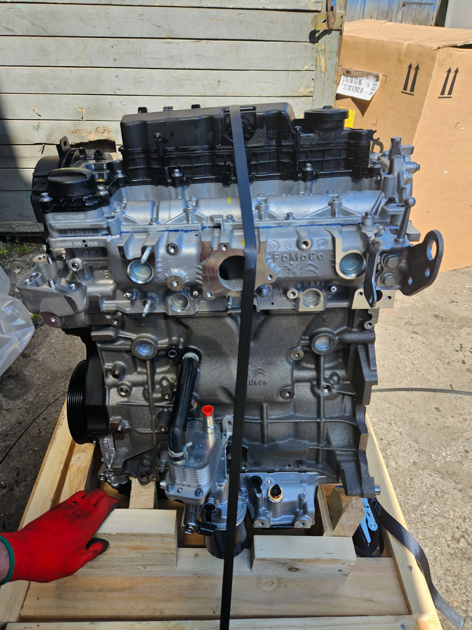 Nowy silnik samochodowy marki FoMoCo, zabezpieczony pasem transportowym na drewnianej palecie, gotowy do montażu lub transportu.