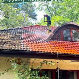 Pracownik w trakcie mycia dachu z czerwonej dachówki za pomocą myjki ciśnieniowej. Widoczna różnica między czystą a zabrudzoną częścią dachu.