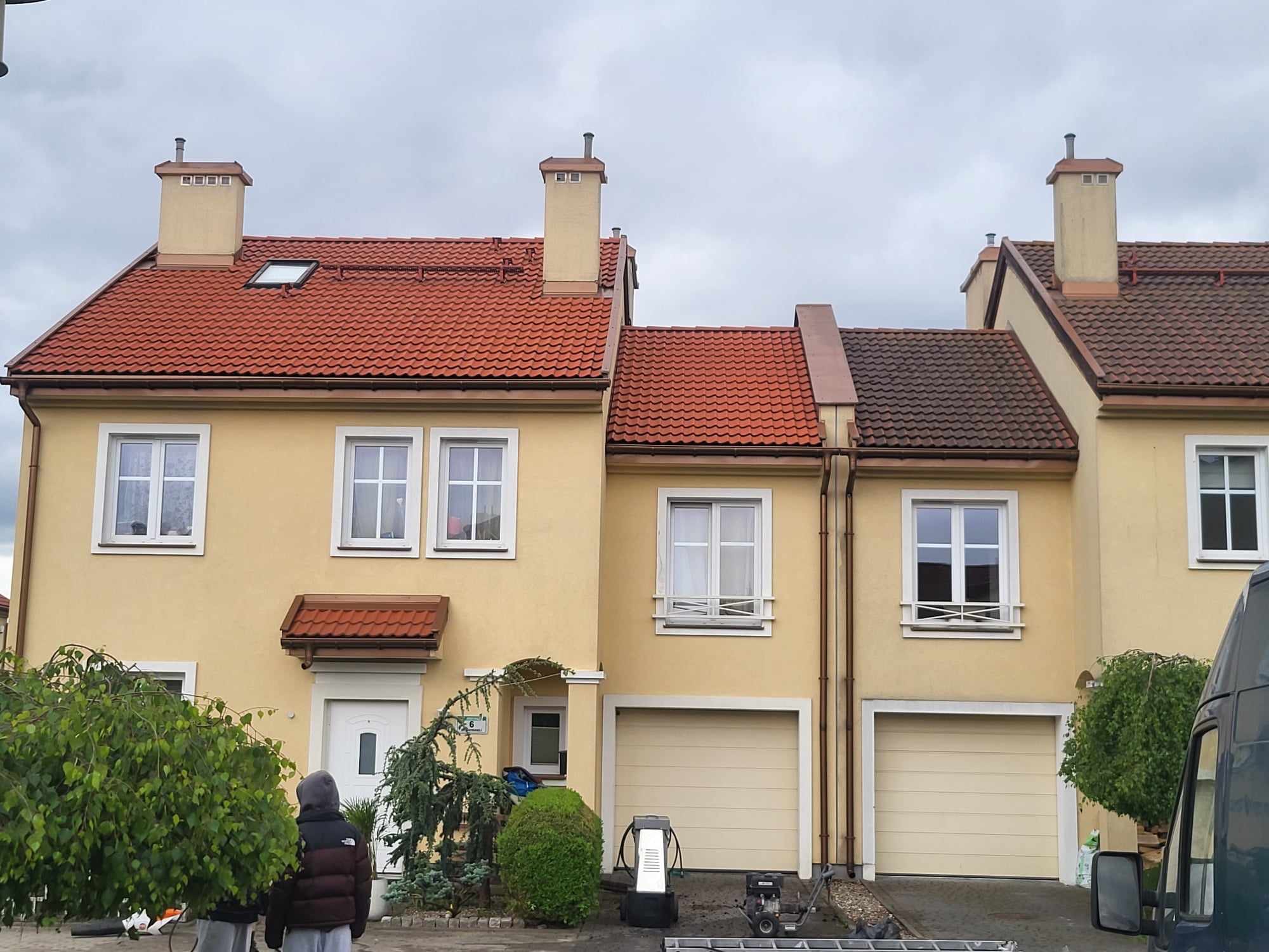 Żółta elewacja szeregowca z czerwoną dachówką, widoczne narzędzia do mycia podjazdu i fragment busa. Usługa mycia elewacji w trakcie realizacji.