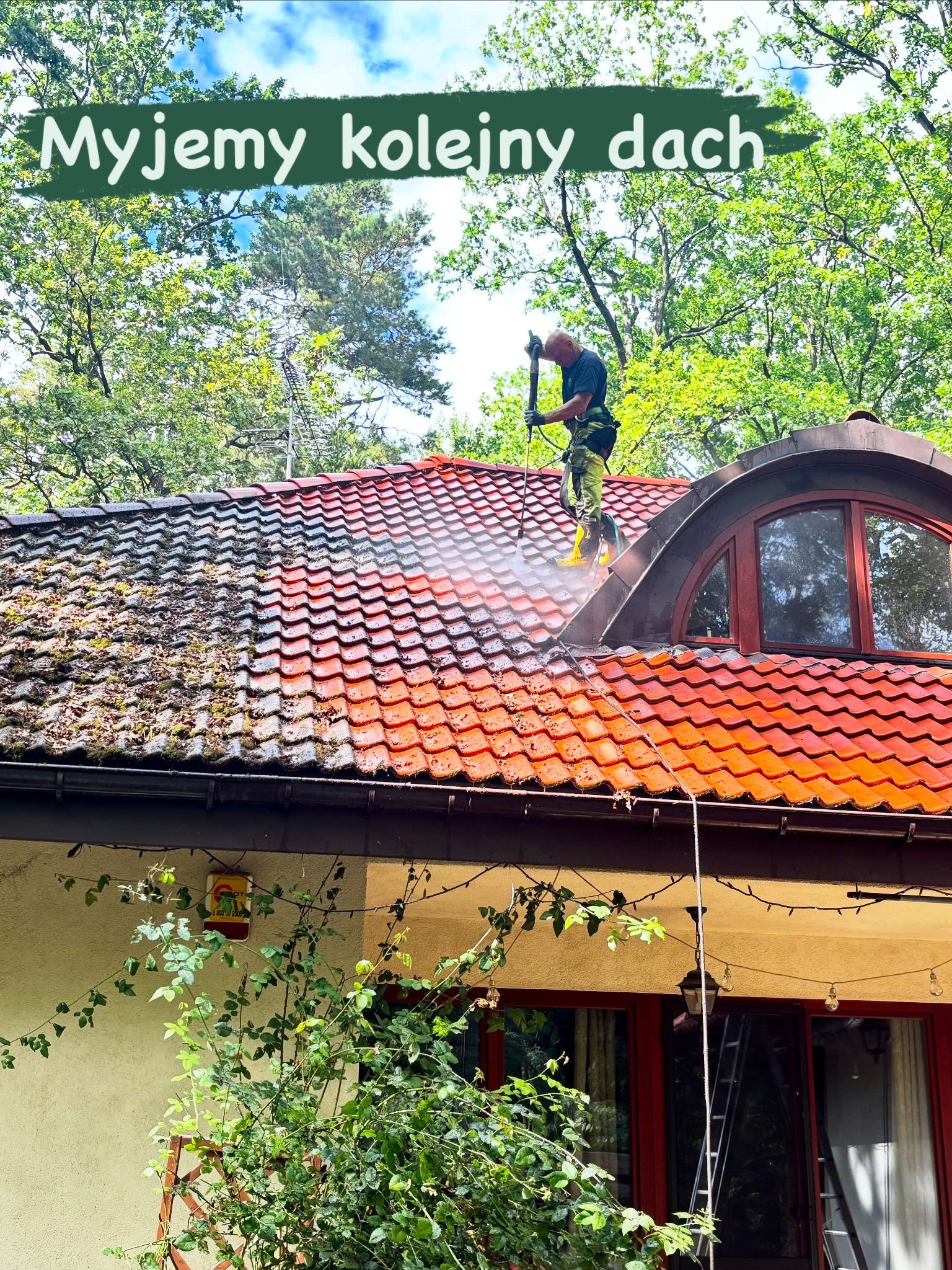 Pracownik w trakcie mycia dachu z czerwonej dachówki za pomocą myjki ciśnieniowej. Widoczna różnica między czystą a zabrudzoną częścią dachu.