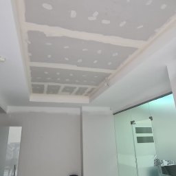 FRB Adrian Martynów - Sufit podwieszany z płyt gipsowo-kartonowych w trakcie montażu, widoczne łączenia płyt i szpachlowanie, klimatyzator na ścianie, przeszklone drzwi w tle.