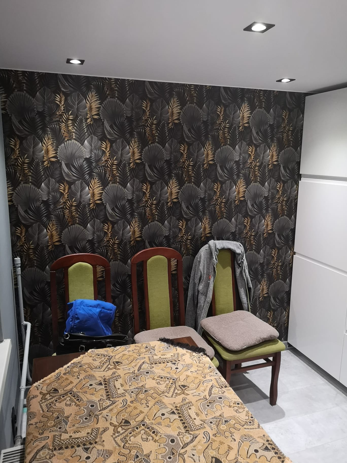 Ściana wykończona tapetą w ciemne liście z elementami złota, trzy krzesła z zielonymi oparciami, na jednym z nich leży szara bluza, a na innym poduszka, widoczne oświetlenie sufitowe