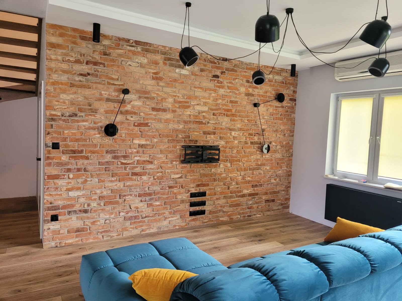 Ceglana ściana w salonie z nowoczesnym oświetleniem, czarnymi włącznikami i gniazdkami, oraz uchwytem na telewizor, widoczny fragment błękitnej sofy.