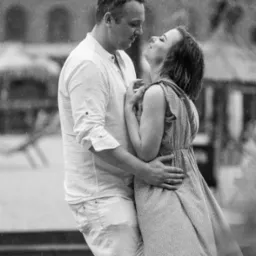 Czarno-białe zdjęcie obejmującej się pary w deszczu, kobieta w kwiecistej sukience z uniesioną głową, mężczyzna w białej koszuli i spodniach, delikatny uśmiech na twarzy kobiety, w tle zamazane...