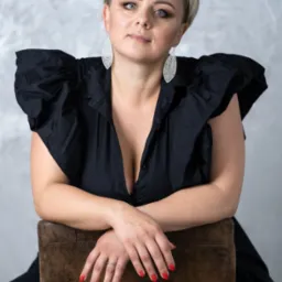 Portret kobiety z krótkimi blond włosami w czarnej sukience z bufkami, opierającej ręce o brązowe krzesło, z widocznym pierścionkiem i czerwonym manicure.