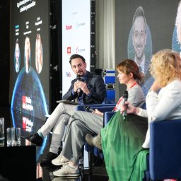 KAROLINA STELLA FLOW PHOTO - Panel dyskusyjny na konferencji: mężczyzna w garniturze mówi do mikrofonu, obok dwie kobiety siedzą w fotelach, w tle ekran z logotypami partnerów i portretami prelegentów.