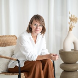 KAROLINA STELLA FLOW PHOTO - Portret kobiety w okularach, siedzącej na wiklinowym krześle z poduszką, w białej koszuli i brązowych spodniach, z dekoracyjnym wazonem z suszem w tle, w jasnym wnętrzu.