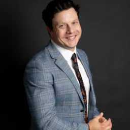 Portret uśmiechniętego mężczyzny w błękitnej marynarce w kratę i wzorzystym krawacie na ciemnym tle, z widocznym zegarkiem na nadgarstku.