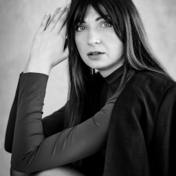 KAROLINA STELLA FLOW PHOTO - Portret kobiety w czarno-białej tonacji, z rękami uniesionymi do twarzy, w ciemnej bluzce i marynarce, z długimi włosami z grzywką, patrzącej w obiektyw.
