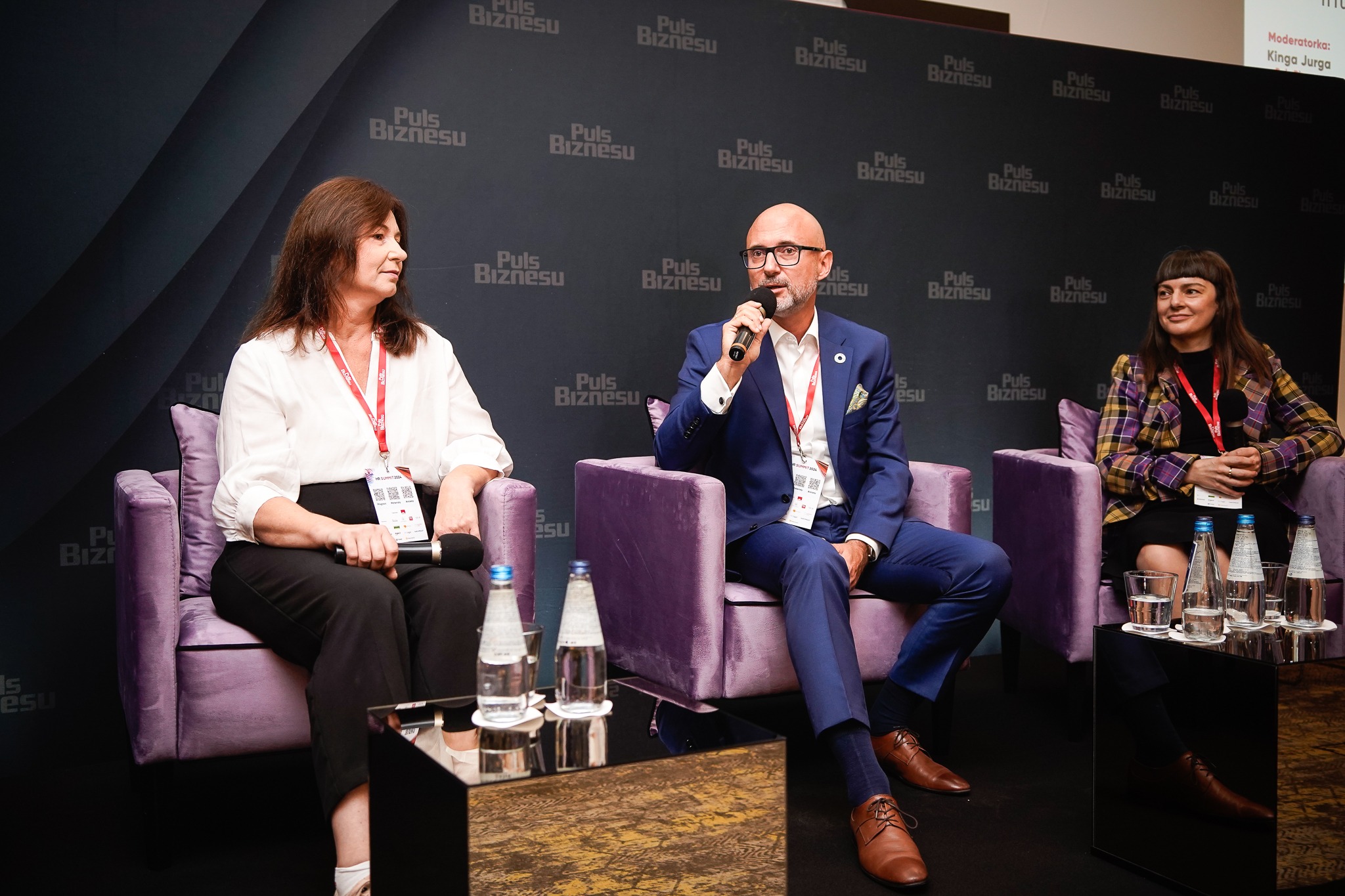 Panel dyskusyjny: troje prelegentów siedzi w fotelach na tle ściany z logo 'Puls Biznesu', mężczyzna w środku mówi do mikrofonu.
