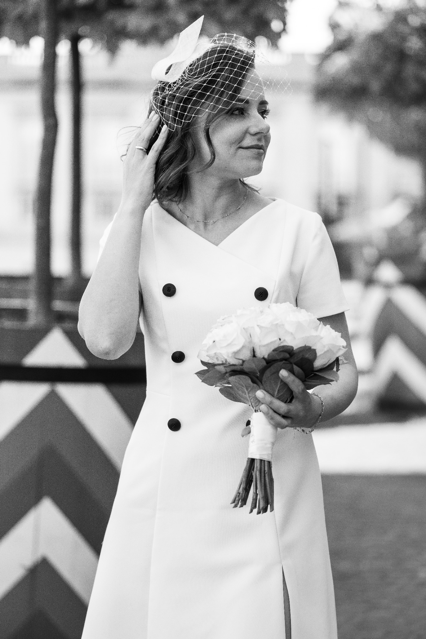 Czarno-białe zdjęcie uśmiechniętej kobiety w białej sukience z dekoracyjnymi guzikami, trzymającej bukiet białych róż i poprawiającej welon z woalką, w tle wzorzysta ściana.