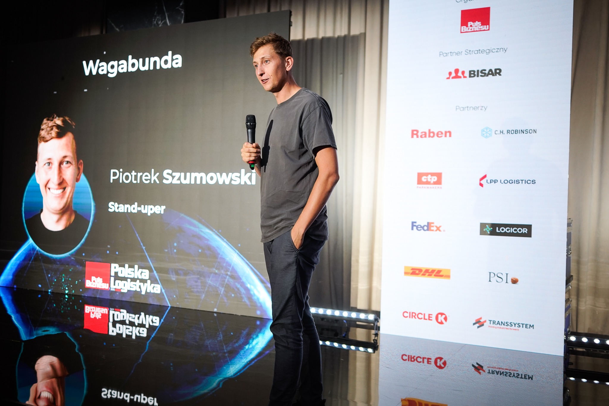 Mężczyzna przemawia do mikrofonu na tle ekranu z napisem 'Wagabunda' i logotypami firm partnerskich.