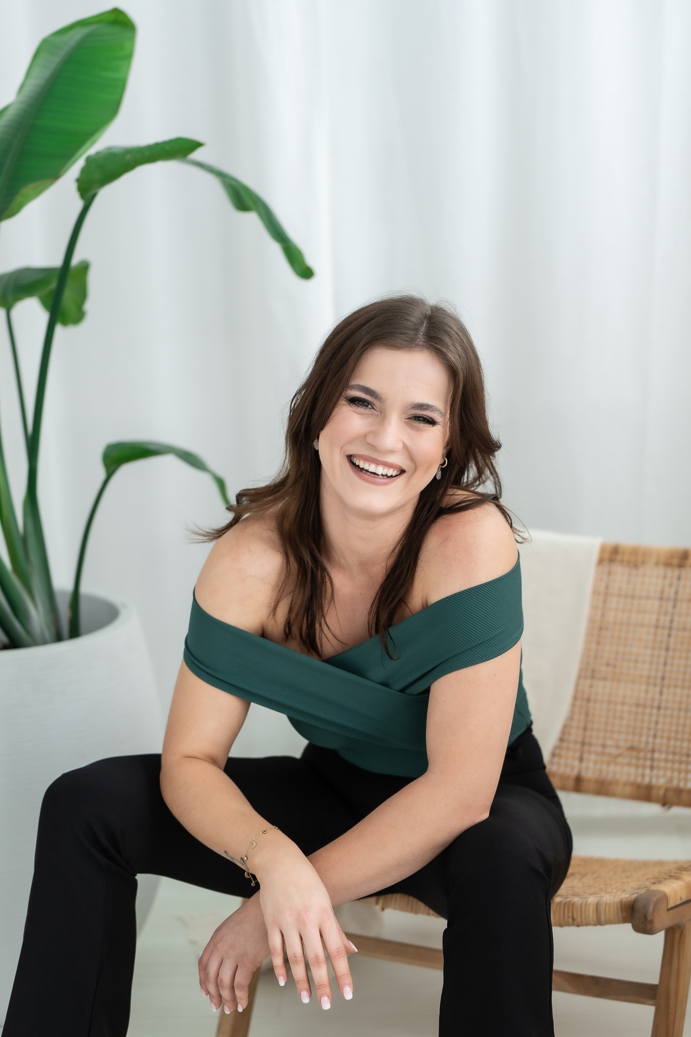 Uśmiechnięta brunetka w zielonej bluzce z odkrytymi ramionami i czarnych spodniach siedzi na wiklinowym krześle, w tle duża roślina w białej donicy i biała zasłona.