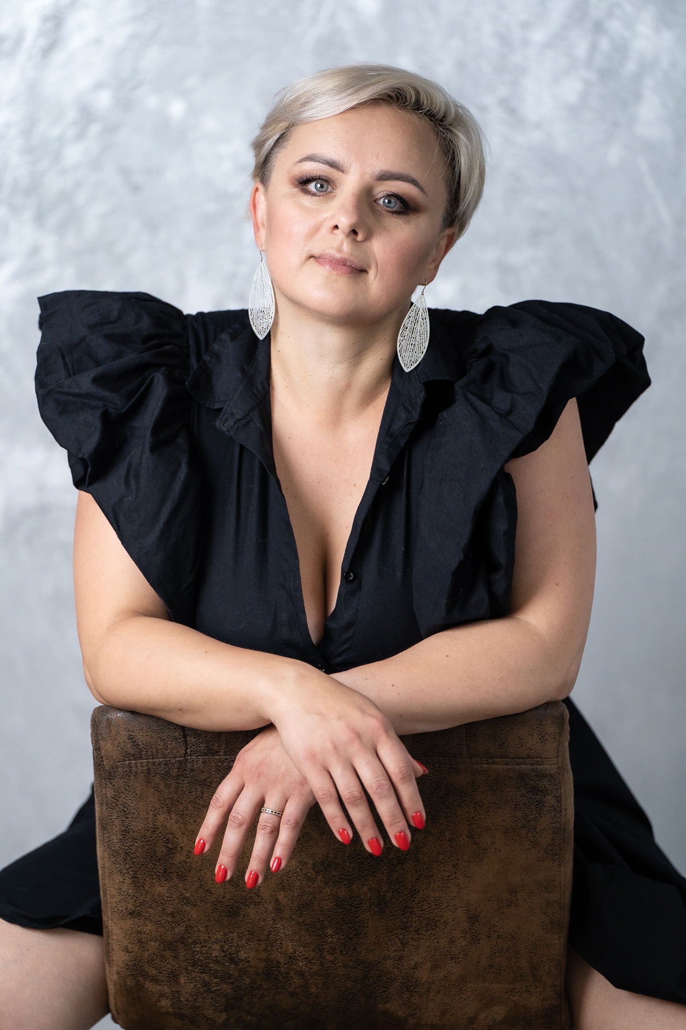 Portret kobiety z krótkimi blond włosami w czarnej sukience z bufkami, opierającej ręce o brązowe krzesło, z widocznym pierścionkiem i czerwonym manicure.