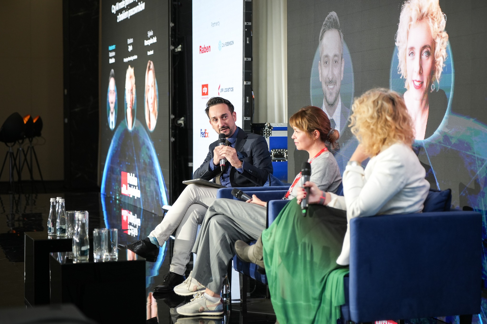 Panel dyskusyjny na konferencji: mężczyzna w garniturze mówi do mikrofonu, obok dwie kobiety siedzą w fotelach, w tle ekran z logotypami partnerów i portretami prelegentów.